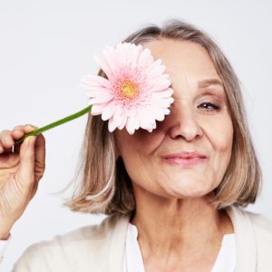 Senior woman managing menopause holding a pink flower to her eye.