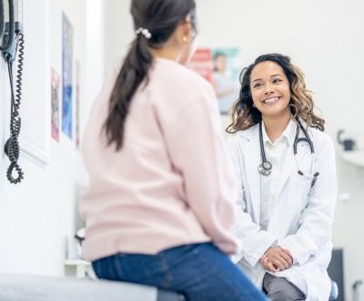 A female doctor wearing a white coat and stethoscope smiling warmly at a patient during an examination room visit.