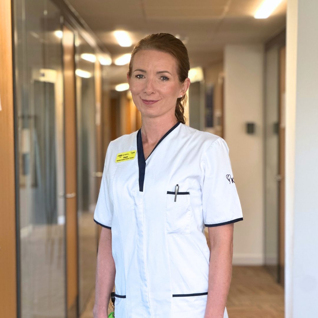 Kasia Rushton a Specialist Inpatient Physical Therapist stands in a hallway, smiling. 