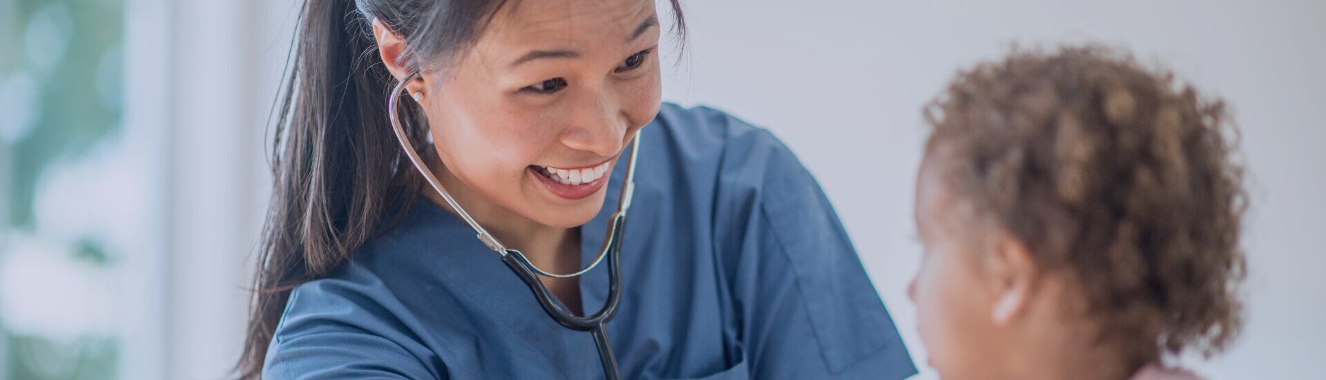 A smiling paediatric clinician in blue scrubs using a stethoscope to examine a young patient at KIMS Hospital.